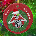 Adorno De Cerámica Skeleton en Navidades de pino fue gracioso para el<br><div class="desc">Este adorno de cerámica colgante tiene un gracioso esqueleto sosteniendo ornamentos con copos de nieve hasta los ojos con un sombrero de Santa roja dentro de una corona de alfiler verde sobre un fondo rojo profundo. Personalizar tu texto en la parte posterior con el y la Feliz Navidad delante con...</div>