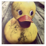 Azulejo ¡Goma amarillo, dudoso, cubierto de tierra!<br><div class="desc">Foto graciosa y burda de un gran pato amarillo de goma sentado en un suelo de madera. Este pato,  tiene un gran pico rojo y grandes ojos mirando fijamente. Cubierta de suciedad de un largo día de diversión y juegos. Parece feliz y juguetón.</div>