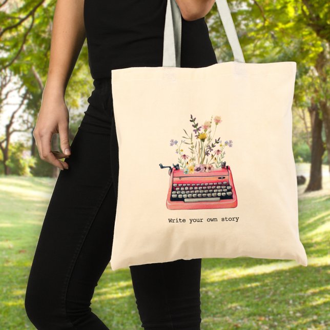 Bolso De Tela Escribe tu propia historia, Escritores (A woman holding a tote bag with a typewriter graphic and wildflowers & text: "write your own story")