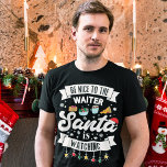 Camiseta Sé amable con el camarero, Santa está mirando con<br><div class="desc">Recuerde a sus clientes que este año permanezcan en la lista con esta divertida y encantadora camiseta de Navidades con la frase "Sé amable con el mesero, Santa está mirando" en tonos negros y modernos, decorada con luces rojas, verdes y amarillas de Navidad, copos de nieve negros, un sombrero de...</div>