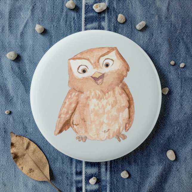 Chapa Redonda De 5 Cm Búho Feliz Acuarela (A watercolor owl cartoon smiling on a pale blue pin, laid on denim amid pebbles and leaves.)