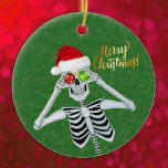 Navidades tontos Gorra de esqueletos Ornamentos Ve<br><div class="desc">Este adorno de cerámica colgante tiene un esqueleto tonto sosteniendo adornos rojos y verdes hasta los ojos mientras sonríe y lleva puesto un gorra rojo de Santa Claus de cuatro pieles sobre un fondo verde con finas motas purpurinoso de oro. Feliz texto navideño en oro en el frente. Personalizar el...</div>