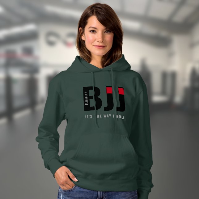 Sudadera Personalizada la BJJ de la mujer (Dark green woman's BJJ hoodie worn by a female model in a gym.)